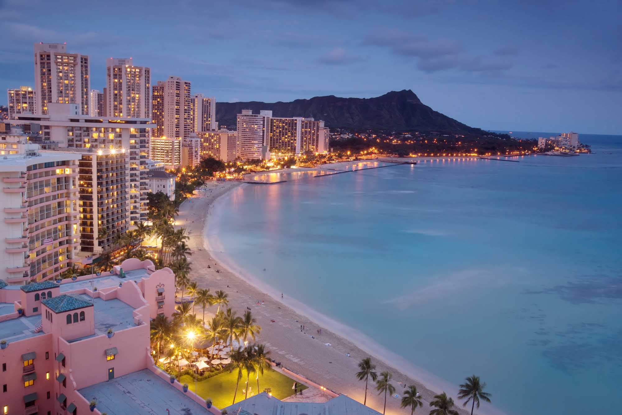 A stunning twilight view of Waikiki Beach, Hawaii, showcasing the iconic crescent shoreline lined with luxury high-rise hotels and resorts illuminated against the deepening blue sky. Diamond Head volcanic crater dominates the background, while palm-lined beaches and calm turquoise waters reflect the warm glow of evening lights along the bustling beachfront.