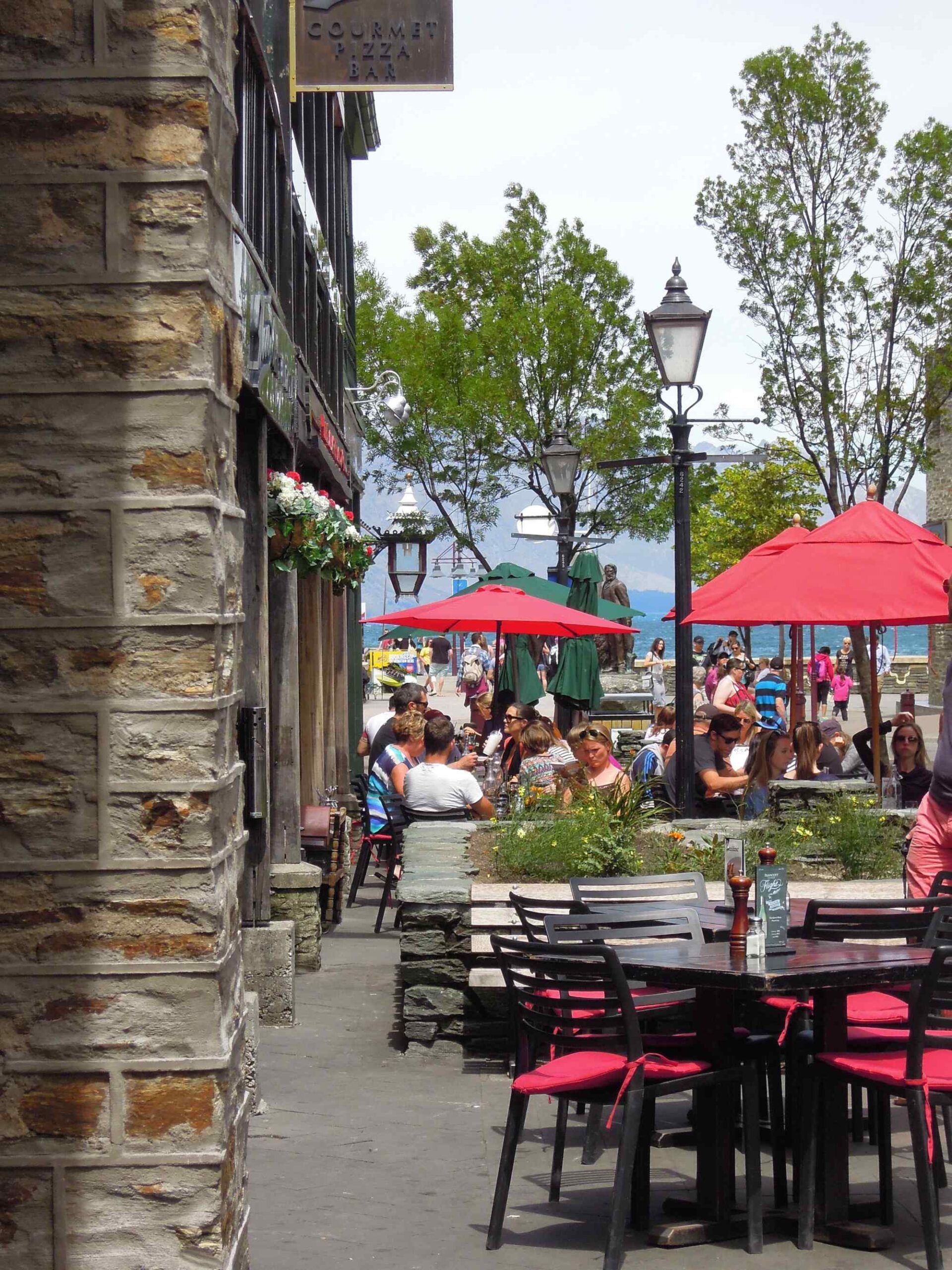 Waterfront Outdoor Dining with Red Umbrellas A bustling waterfront dining area features black tables with red cushioned chairs under bright red umbrellas, with a stone building featuring a gourmet bar sign on the left. In the background, a scenic beach with mountains is visible, populated with beachgoers and additional red umbrellas creating a vibrant summer atmosphere.