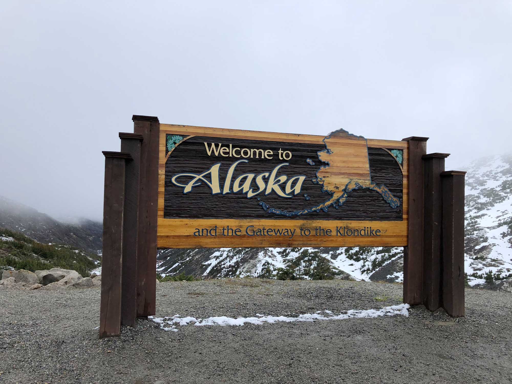 A rustic wooden welcome sign marking the entrance to Alaska, identifying it as the gateway to the Klondike. The sign features carved text and the state outline against a backdrop of snow-capped mountains and misty valleys.