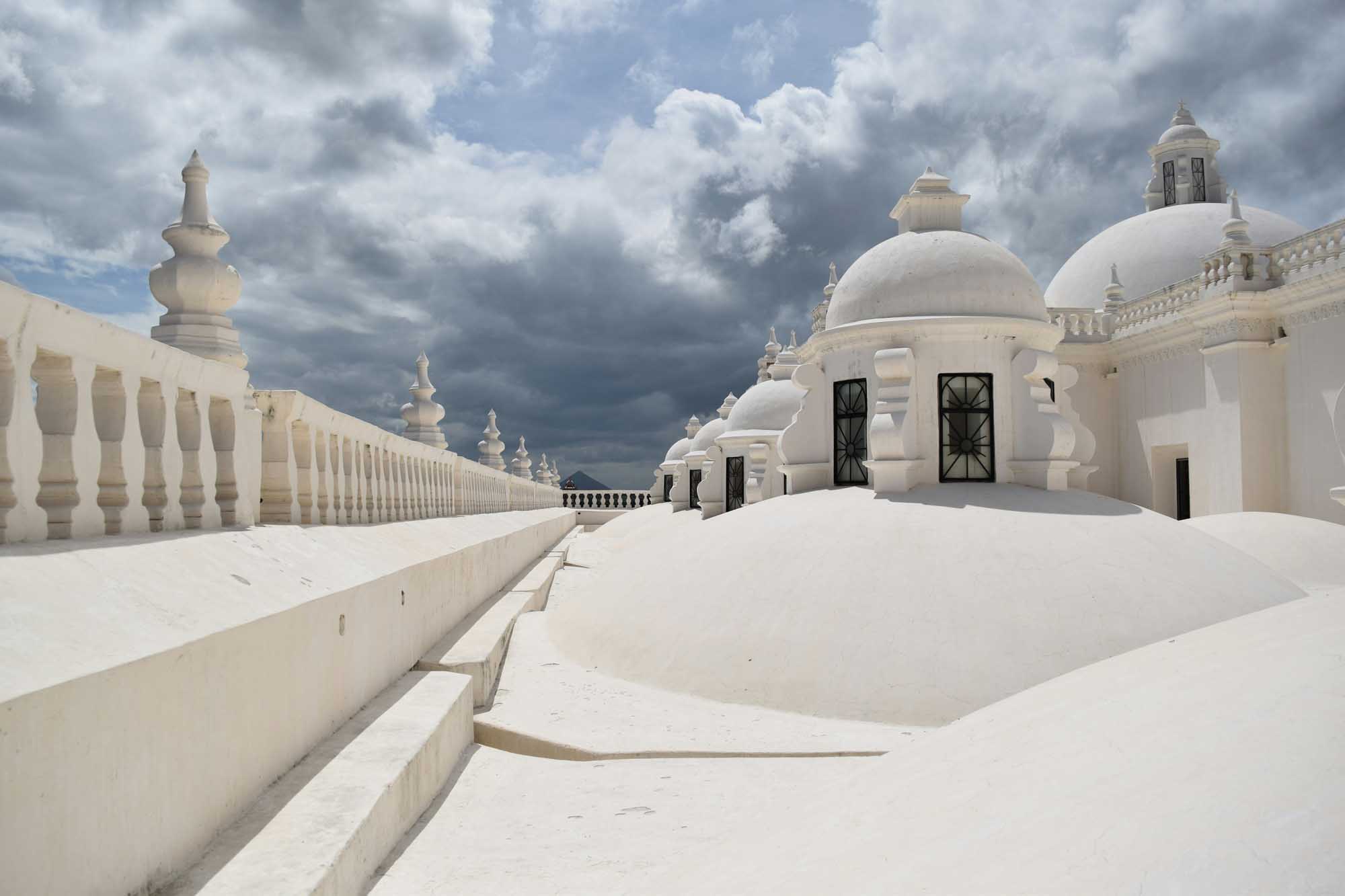 A stunning rooftop terrace featuring pristine white-washed architecture with iconic domed structures and ornamental balustrades, characteristic of Mediterranean or Middle Eastern design. The dramatic cloudy sky creates a striking contrast against the brilliant white buildings adorned with decorative finials and geometric details.