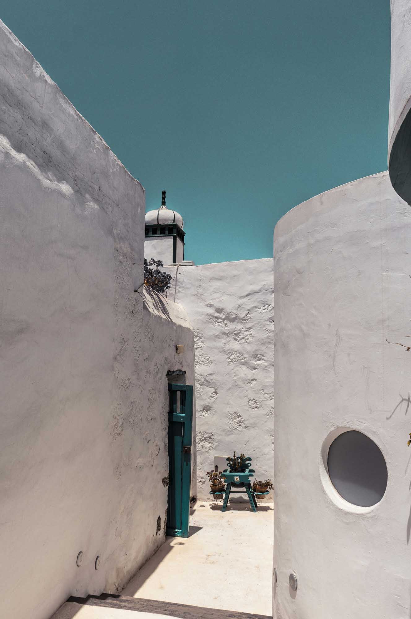 A narrow, whitewashed alleyway framed by pristine white-washed walls features a traditional minaret tower with a distinctive dome visible in the center background. A turquoise door and circular window accent the Mediterranean or North African architectural style, with a clear turquoise sky providing striking contrast to the white structures.