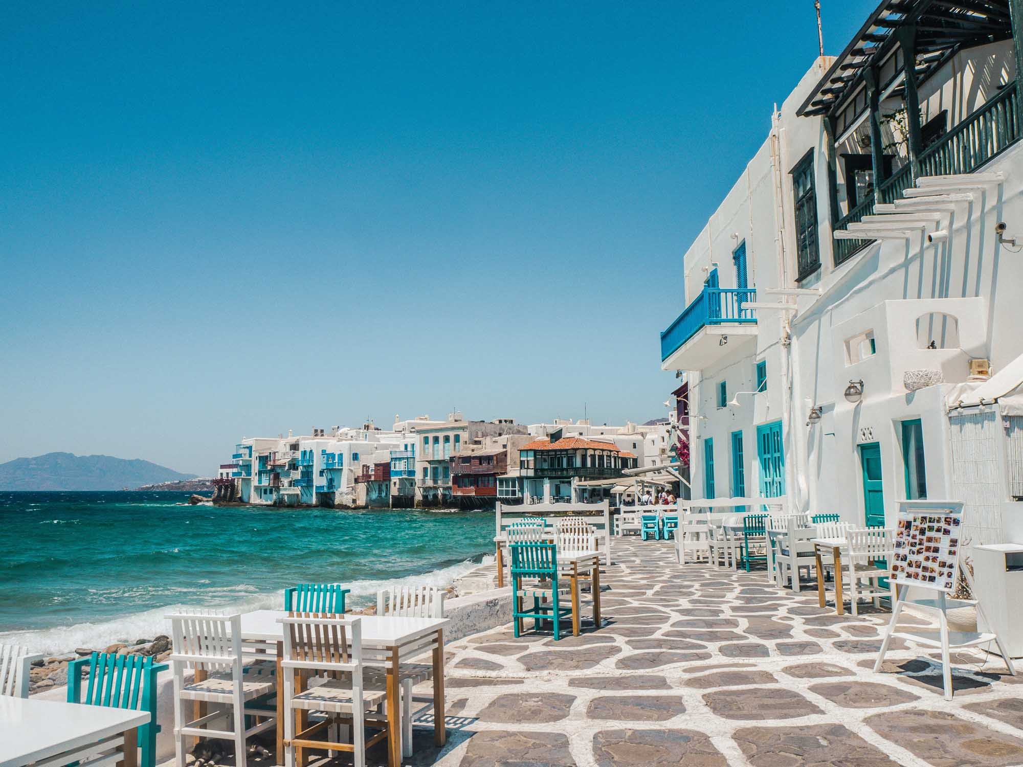 A picturesque Greek island waterfront scene featuring whitewashed buildings with turquoise accents and wooden chairs arranged on a traditional patterned stone plaza overlooking crystal-clear turquoise waters. Charming architecture typical of the Cycladic islands lines the waterfront, creating an idyllic Mediterranean coastal setting.