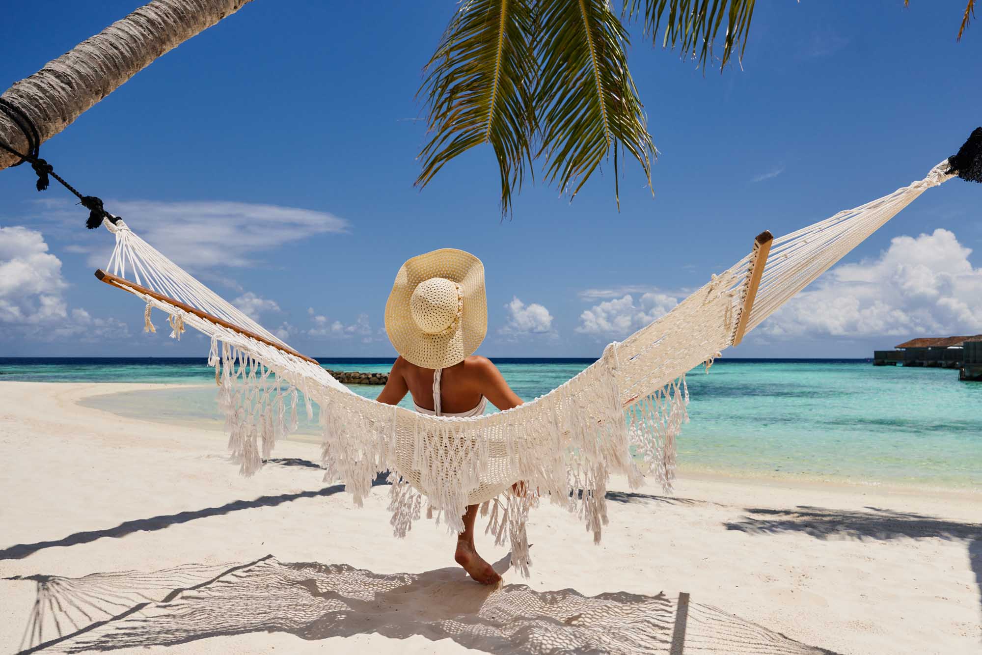 A rear view of a woman wearing a straw sun hat relaxing in a white macramé hammock strung between palm trees on a pristine tropical beach, overlooking turquoise waters and a clear blue sky dotted with white clouds.